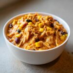A close-up of a white bowl filled with creamy, shredded slow cooker buffalo chicken chili topped with corn and black beans.