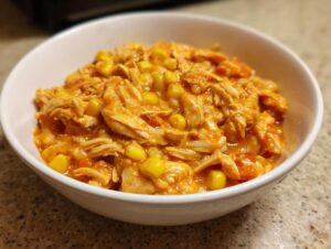 A white bowl filled with shredded chicken and bright yellow corn kernels in a creamy, orange sauce, representing slow cooker buffalo chicken chili.