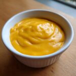 A close-up of thick, vibrant orange nacho cheese sauce swirled inside a small white ceramic ramekin.