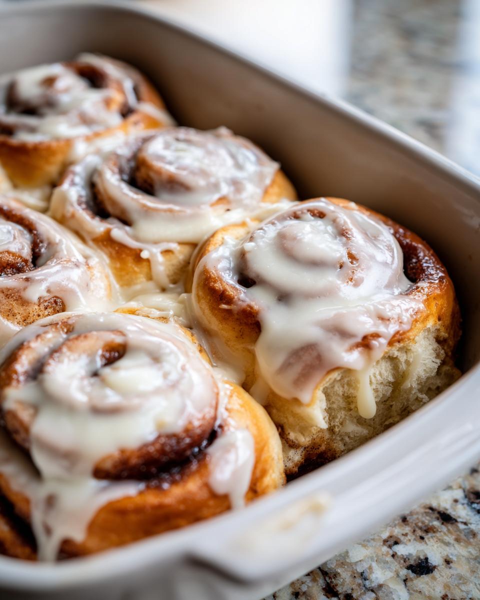 Close-up of soft, golden brown cinnamon buns topped with thick white icing in a baking dish.