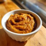 A close-up of thick, rich, orange-brown pear butter swirled in a small white bowl, sitting on a wooden surface.
