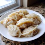 A white plate holding several freshly steamed Traditional Chinese Dumplings with crimped edges, sitting on a granite countertop.