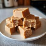 A close-up stack of creamy, light brown peanut butter fudge squares piled on a small white plate.