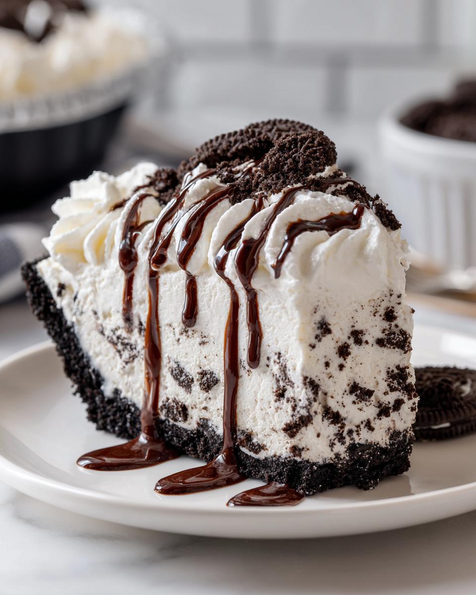 A decadent slice of no-bake ice cream pie featuring a dark cookie crust, creamy filling with cookie chunks, topped with whipped cream and chocolate syrup.