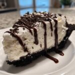 A decadent slice of no-bake ice cream pie featuring a dark crust, creamy white filling, chocolate syrup drizzle, and cookie crumbs.