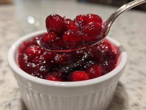 A spoonful of glistening, whole-berry cranberry sauce being lifted from a white ramekin.