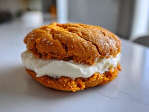 A close-up of a soft, orange pumpkin cookie sandwich filled with thick cream cheese frosting.