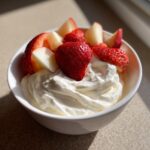 A white bowl filled with creamy fruit dip, topped with fresh strawberries and apple slices.