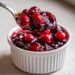 A spoonful of glistening, whole-berry cranberry sauce being lifted from a white ramekin.
