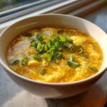 A close-up of a bowl of hot egg drop soup, featuring wispy yellow egg ribbons garnished with fresh green scallions.