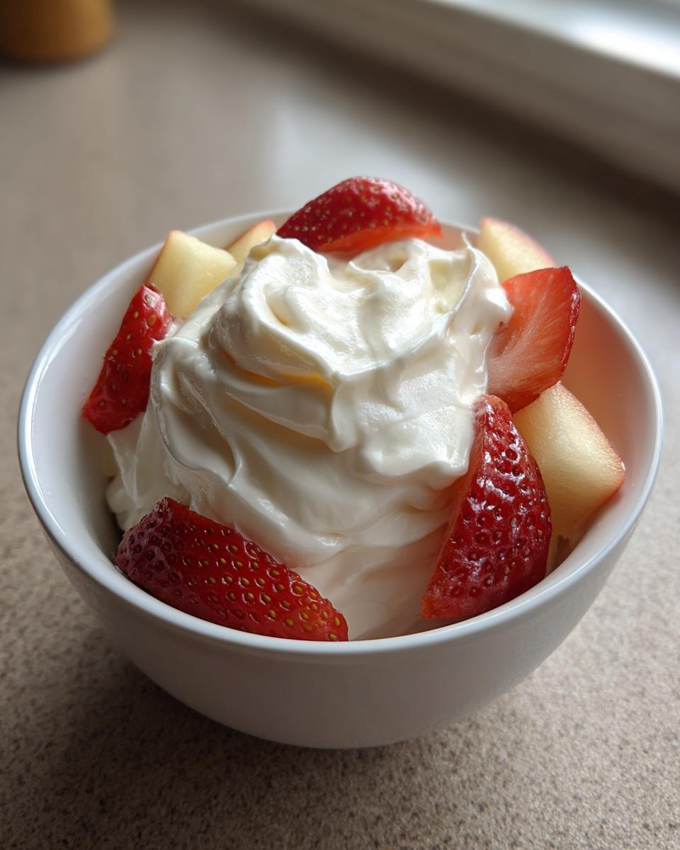 A white bowl filled with creamy fruit dip topped with fresh sliced strawberries and apple pieces.