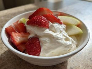 Close-up of a white bowl filled with creamy fruit dip, topped with sliced strawberries and apple wedges.