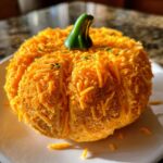 A festive pumpkin cheese ball covered entirely in shredded cheddar cheese with a green stem on top.