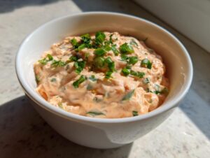 Close-up of creamy, orange-hued remoulade sauce topped with fresh chopped green onions in a white bowl.