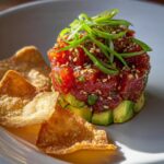 Close-up of a perfectly stacked tuna tartare tower layered over avocado, topped with scallions and sesame seeds, served with crispy wonton chips.