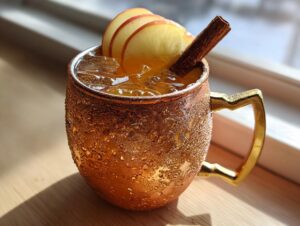 A refreshing apple cider moscow mule served in a frosty copper mug, garnished with apple slices and a cinnamon stick.