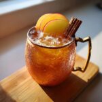 A chilled apple cider moscow mule garnished with apple slices and a cinnamon stick in a textured amber mug.