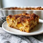A square slice of apple cinnamon baked oatmeal with a dark, sugary crumb topping, served on a white plate.
