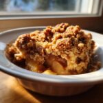 Close-up of a warm serving of apple crisp recipe in a white bowl, featuring chunky baked apples and a golden oat topping.