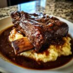 A close-up of a tender braised short ribs piece, coated in rich sauce, served over creamy mashed potatoes.