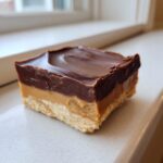 A single square of rich buckeye fudge showing distinct layers of crust, peanut butter filling, and chocolate topping.