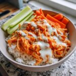 A bowl filled with white rice topped with shredded buffalo chicken and drizzled with ranch dressing, served with celery and carrot sticks.