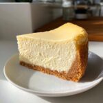 A thick, creamy slice of classic cheesecake with a golden graham cracker crust, served on a white plate.