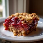 A close-up of a square slice of cherry dump cake showing the bright red cherry filling and crumbly topping on a white plate.