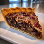 Close-up of a rich slice of chocolate bourbon pecan pie showing the flaky crust and gooey filling topped with pecans.