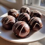A plate of homemade chocolate covered oreo balls drizzled with white chocolate, catching sunlight.
