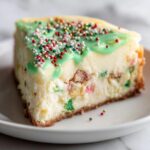 A close-up of a slice of Christmas tree cheesecake topped with bright green icing and festive red, white, and gold sprinkles.