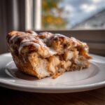 A close-up of a warm slice of cinnamon roll apple pie topped with white icing on a white plate.