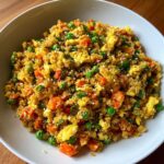 A close-up of a white bowl filled with vibrant cauliflower fried rice, mixed with scrambled egg, peas, carrots, and sesame seeds.