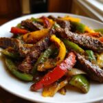 Close-up of sizzling steak fajitas featuring strips of seasoned steak mixed with red, yellow, and green bell peppers.