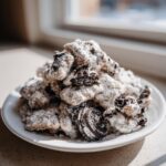 A mound of cookies and cream puppy chow dusted heavily with powdered sugar, featuring visible Oreo pieces.