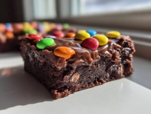 A close-up, sunlit shot of a fudgy square of cosmic brownies topped with thick chocolate frosting and colorful candy pieces.