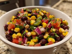 Close-up of colorful cowboy caviar featuring black beans, corn, tomatoes, and red onion in a white bowl.
