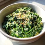 Close-up of rich creamed spinach recipe served in a white bowl, topped with grated cheese and spices.