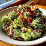 A close-up of a bowl of creamy broccoli salad topped with crispy bacon bits and dried cranberries.