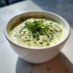 Close-up of creamy dill pickle dip in a white bowl, topped generously with fresh dill and chives.