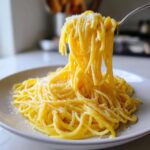 A fork lifts a generous portion of bright yellow, creamy lemon pasta topped with grated Parmesan cheese.