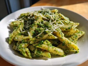 A close-up shot of penne pasta coated in vibrant green sauce, served in a white bowl, topped with shredded Parmesan cheese for creamy pesto pasta.