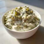 Close-up of a white bowl filled with creamy, chunky pickle dip mixed with fresh dill.