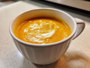 Close-up of rich orange pumpkin soup garnished with a swirl of cream and black pepper in a white mug.