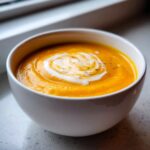 A close-up of a white bowl filled with smooth, vibrant orange pumpkin soup, swirled with cream and pepper.