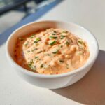 Close-up of creamy, orange-hued remoulade sauce topped with fresh chopped chives in a small white bowl.