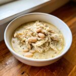 A close-up of a white bowl filled with creamy white chicken chili, featuring shredded chicken and white beans.