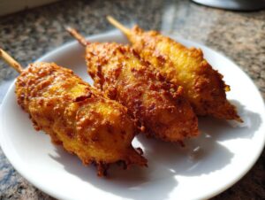 Three golden brown, freshly fried corn dogs on wooden sticks resting on a white plate.