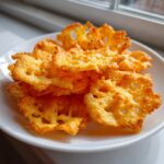 A pile of golden, crispy, baked cheese crackers with lacy edges served on a white plate near a window.