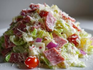 A close-up view of a vibrant grinder salad piled high with crisp lettuce, salami, cheese cubes, red onion, and cherry tomatoes, all coated in dressing.